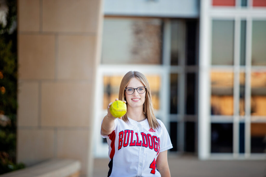 Plainview Bulldog softball player standing in front of the highschool holding a softball