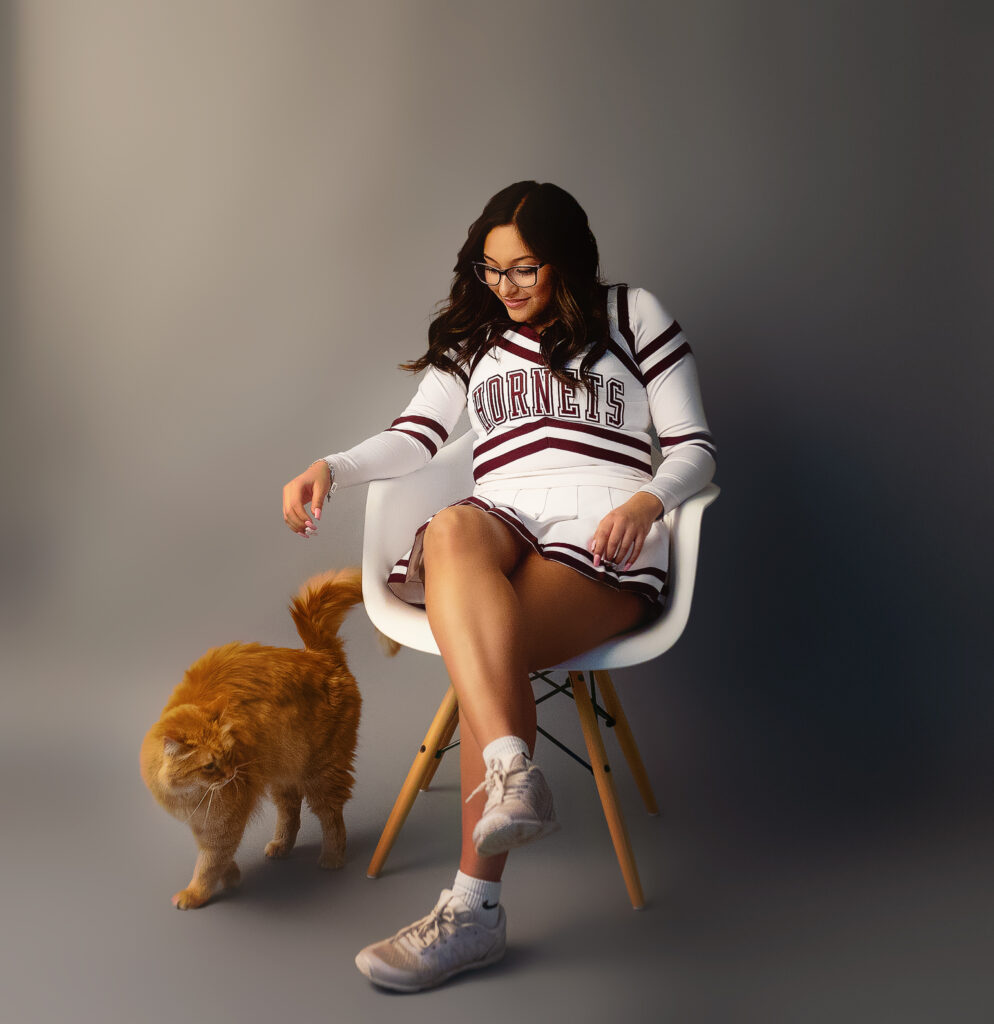 Varsity cheerleader sitting in white chair with an orange cat at her feet