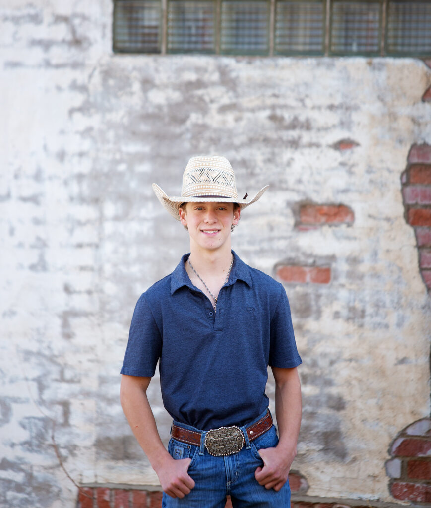 Senior boy in blue shirt standing against a brick wall with a straw cowboy hat 